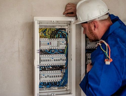 An electrician in a hard hat and blue uniform works on a complex and numerous wires and switches