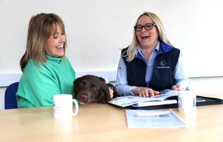 Jeanette and Mandy laughing at a desk with the dog interrupting